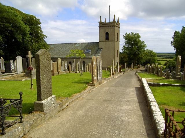 A churchyard with a prominent tower, rows of headstones, and a clear sky.