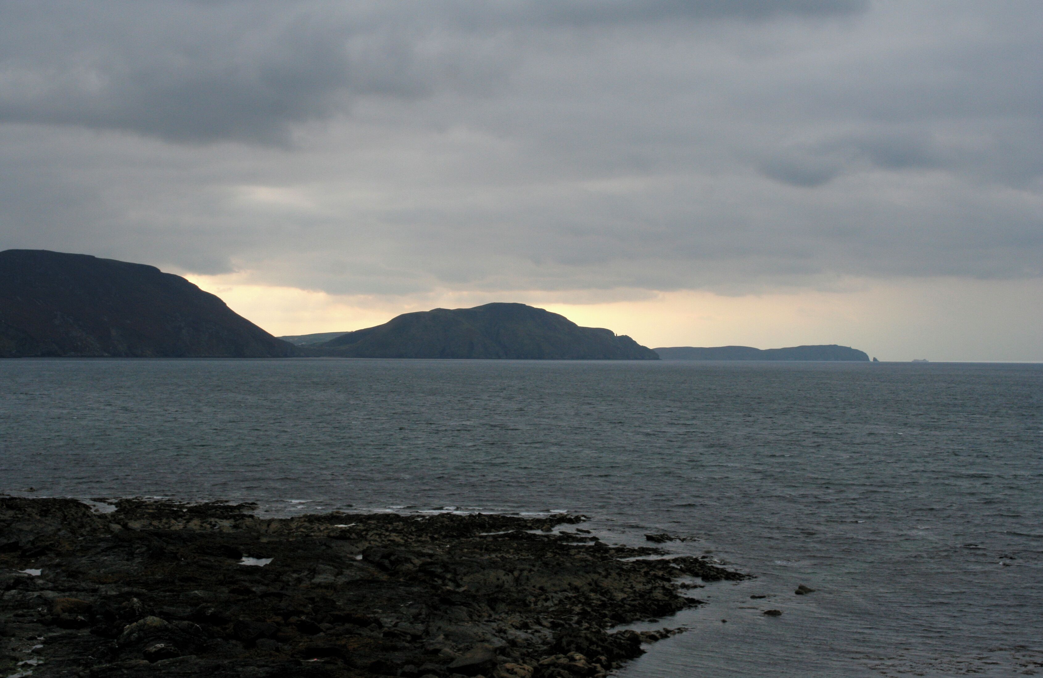 A rocky shoreline with calm water and distant mountains under a cloudy sky.