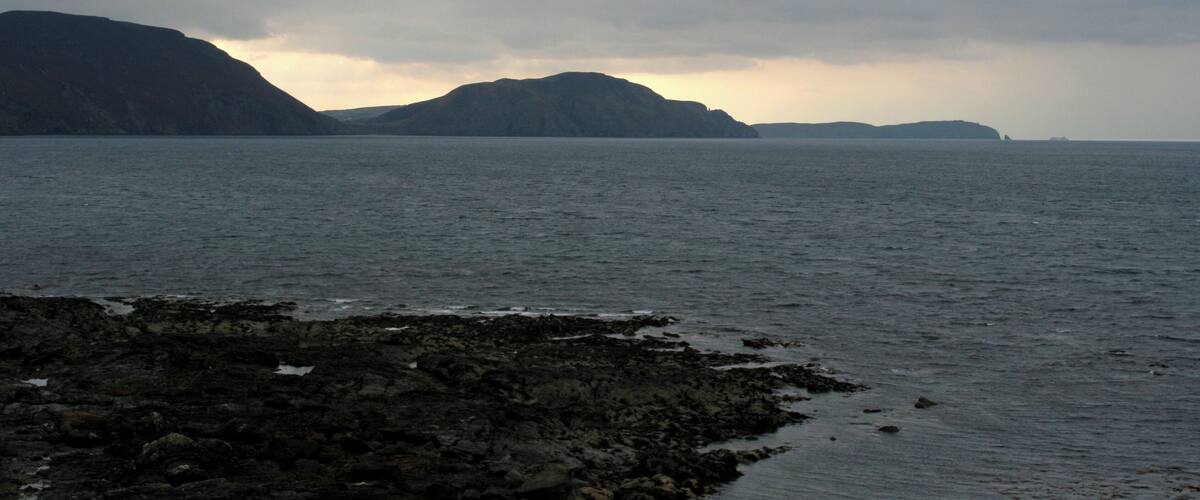 A rocky shoreline with calm water and distant mountains under a cloudy sky.
