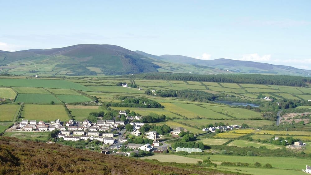 Photo showing the varied terrain (mountains, hills, plains) and land uses (residential, farming, water reservoir) of Isle of Man. This view of the south central portion of the island includes the town of Foxdale, Mines Road, Kionslieau Road, and Kionslieau Reservoir. The camera is located along the A36 Road oriented toward the northeast. Foxdale is a village in the parish of Patrick.