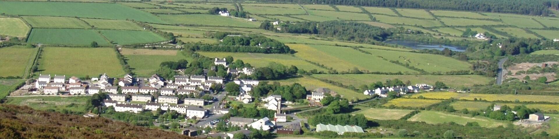 Photo showing the varied terrain (mountains, hills, plains) and land uses (residential, farming, water reservoir) of Isle of Man. This view of the south central portion of the island includes the town of Foxdale, Mines Road, Kionslieau Road, and Kionslieau Reservoir. The camera is located along the A36 Road oriented toward the northeast. Foxdale is a village in the parish of Patrick.