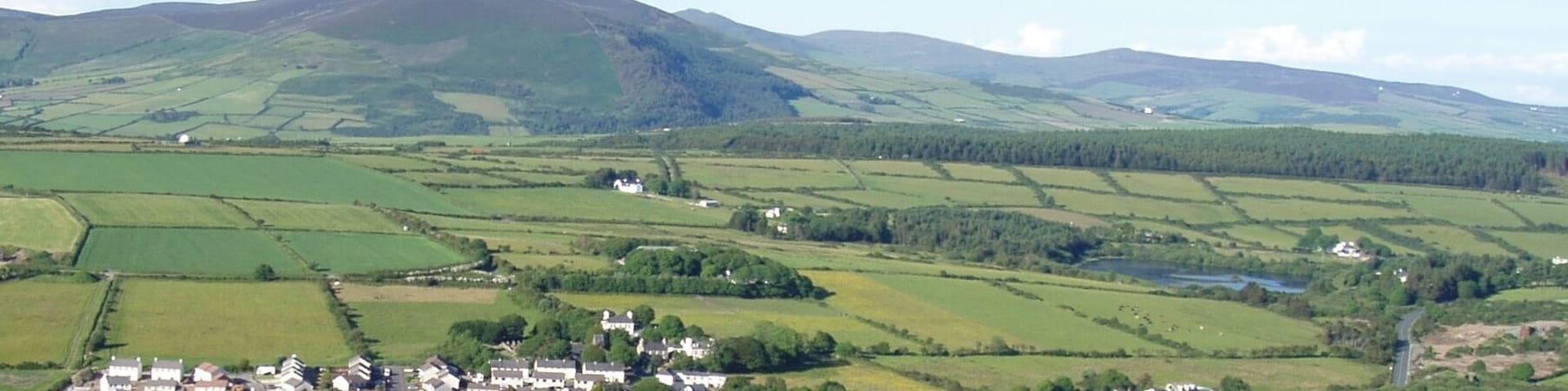 Photo showing the varied terrain (mountains, hills, plains) and land uses (residential, farming, water reservoir) of Isle of Man. This view of the south central portion of the island includes the town of Foxdale, Mines Road, Kionslieau Road, and Kionslieau Reservoir. The camera is located along the A36 Road oriented toward the northeast. Foxdale is a village in the parish of Patrick.