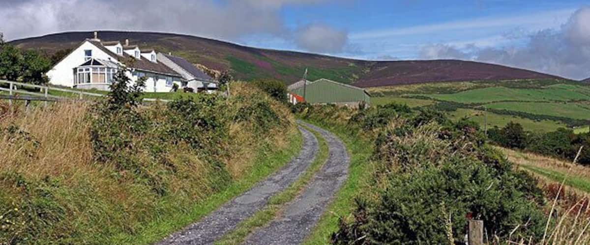 A rural landscape with a white house, a winding road, and rolling hills under a partly cloudy sky.