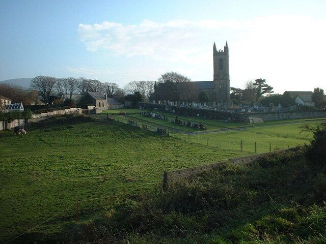 A grassy field with a stone wall, a church with a tall tower, and a few buildings in the background.