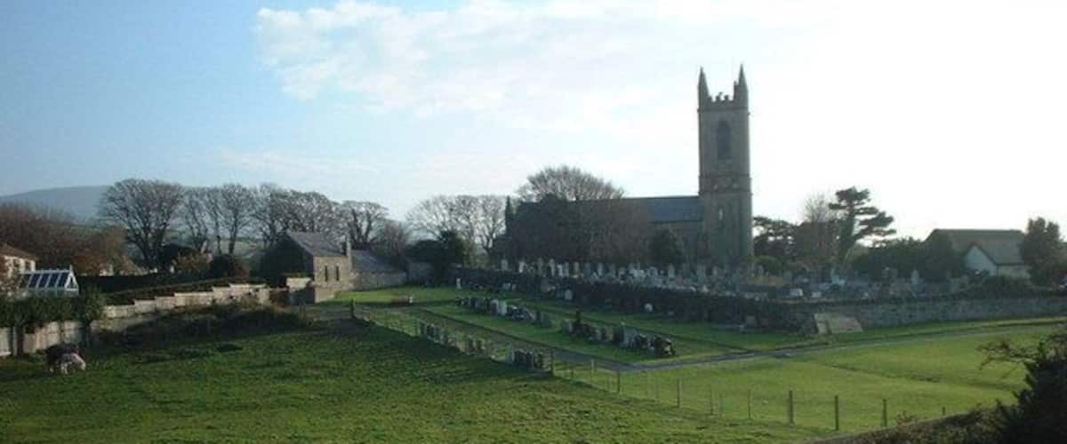A grassy field with a stone wall, a church with a tall tower, and a few buildings in the background.