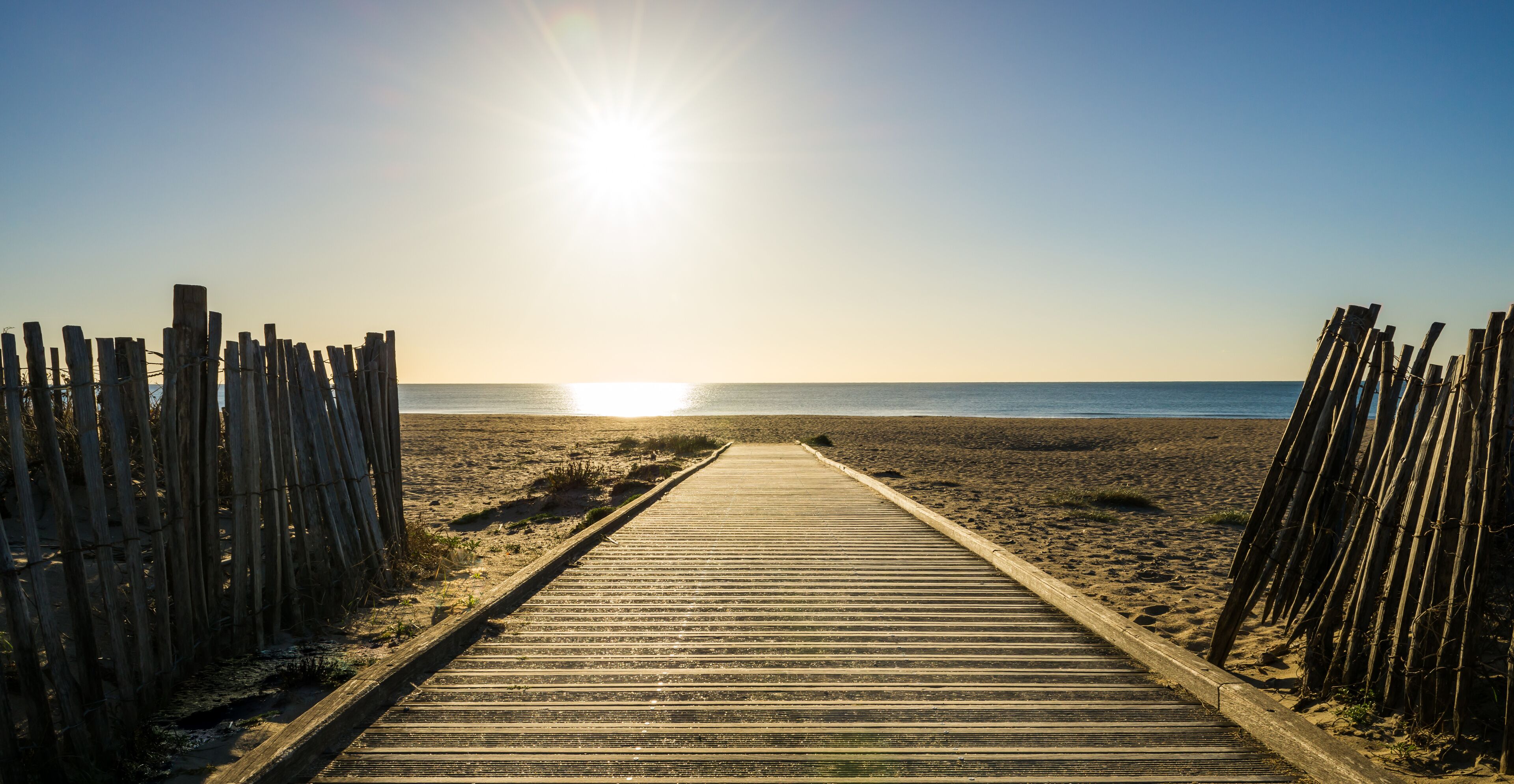 A wooden boardwalk leading to a beach with a clear sky and the sun shining.