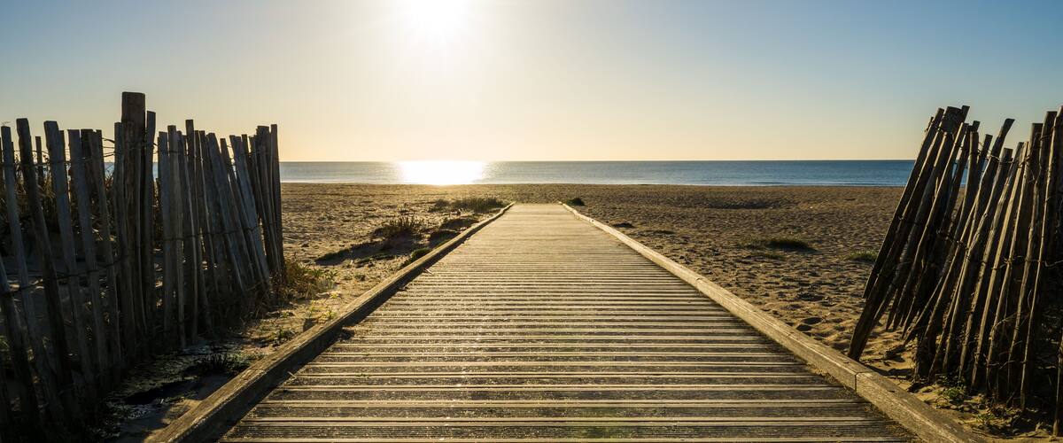 A wooden boardwalk leading to a beach with a clear sky and the sun shining.