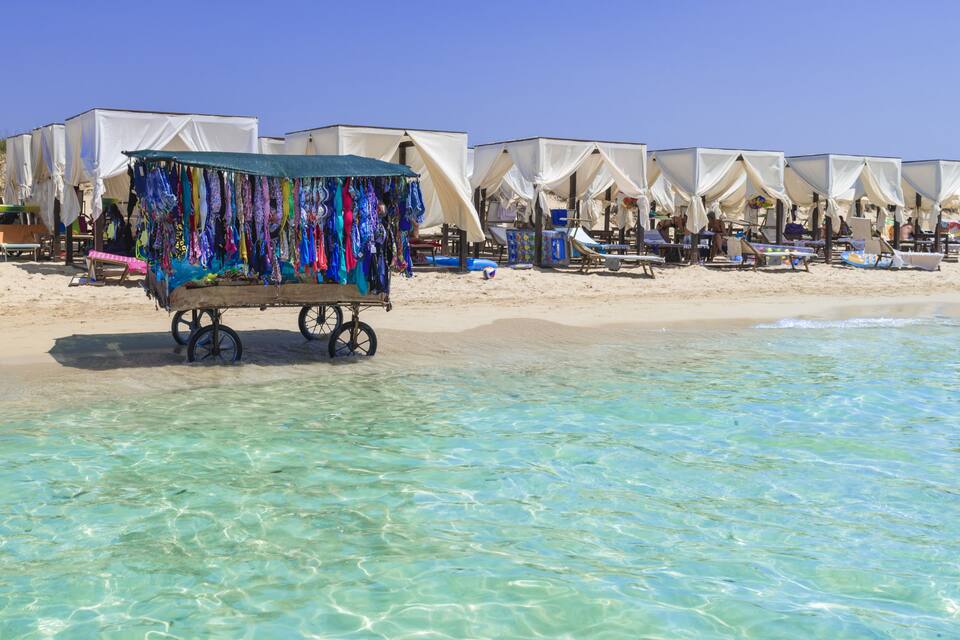 Een strand met helder turquoise water, witte tenten en een kar vol kleurrijke stoffen.