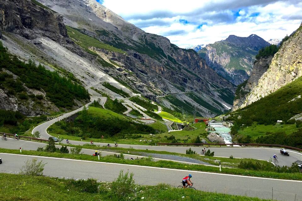 Eine Bergstraße schlängelt sich durch ein Tal, darunter fahren Radfahrer und fließt ein Fluss.
