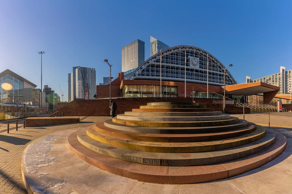 A city square with a circular monument inscribed with names, surrounded by modern buildings and a tram.