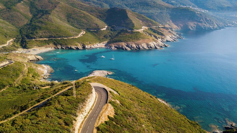 A coastal road winding through green hills with a beach and boats in the background.