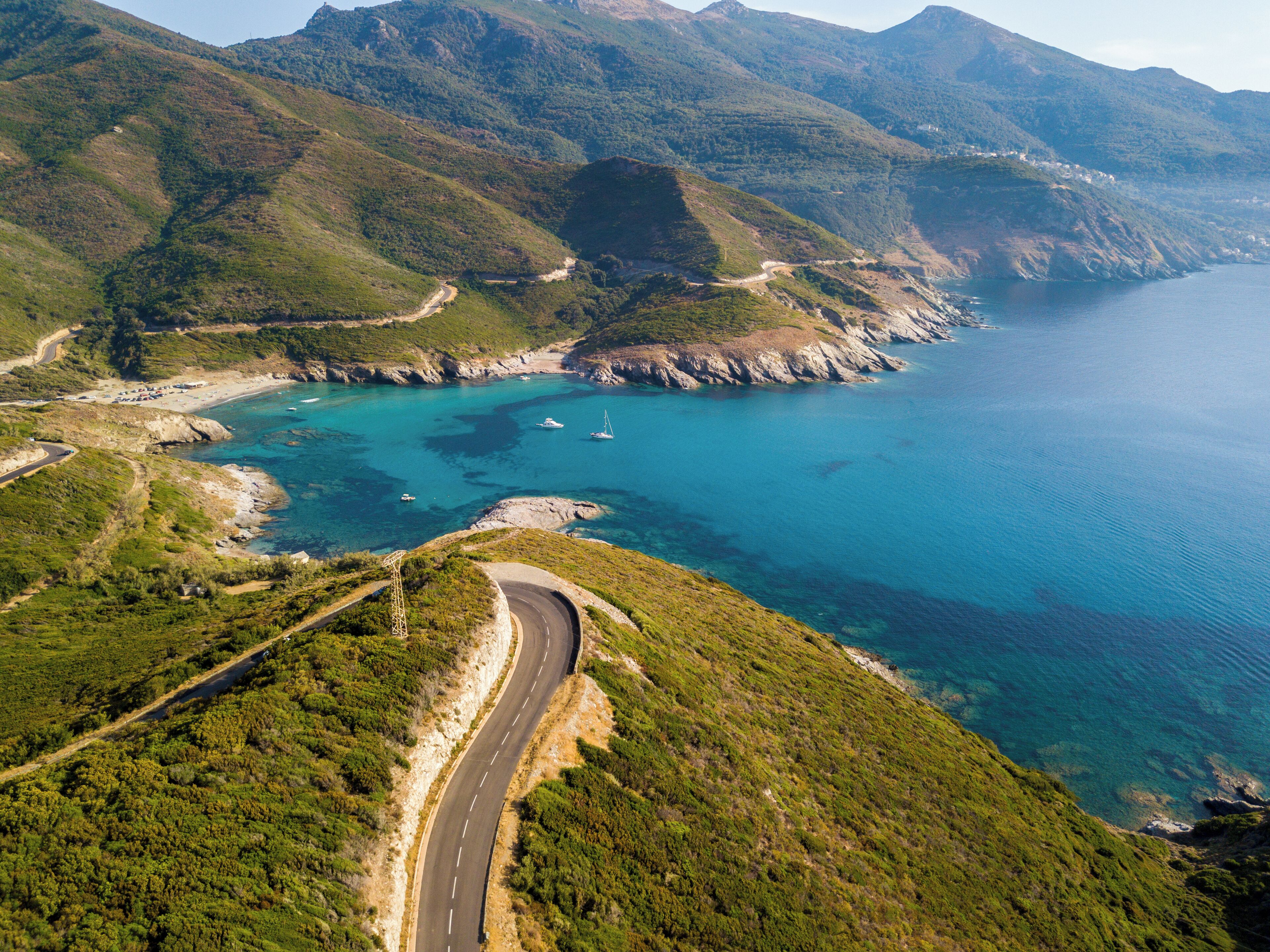 A coastal road winding through green hills with a beach and boats in the background.