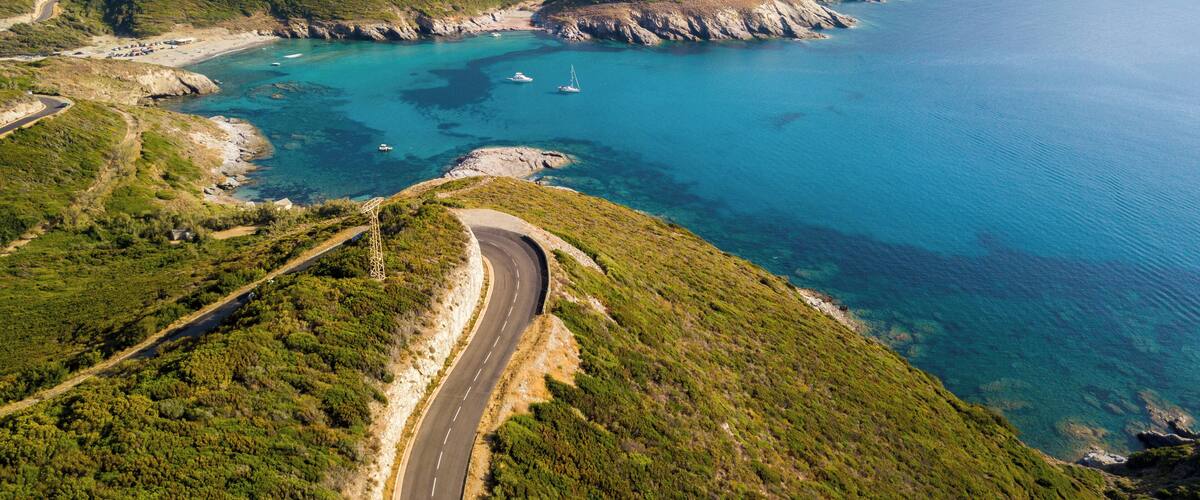 A coastal road winding through green hills with a beach and boats in the background.