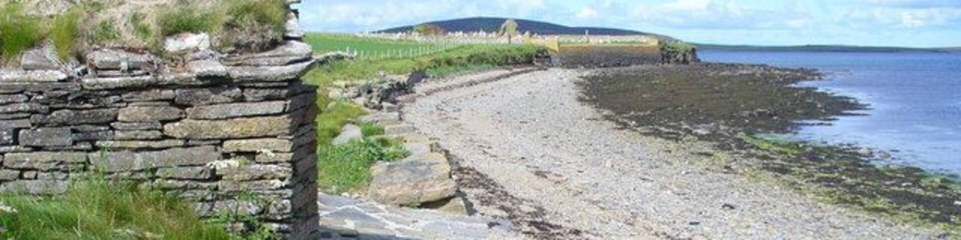 Noust at South Aittit Drystane boathouse on the shore of Bay of Hinderayre. In the distance is the ruined kirk and cemetery. The dark hill on the skyline is on Gairsay.