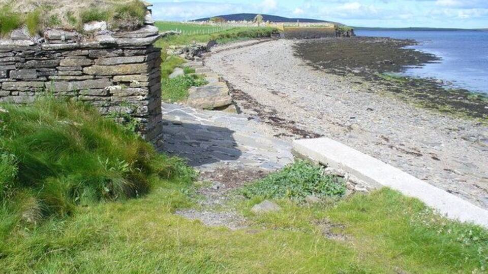 A coastal stone wall, grassy area, and a curved stone pathway leading to a beach.