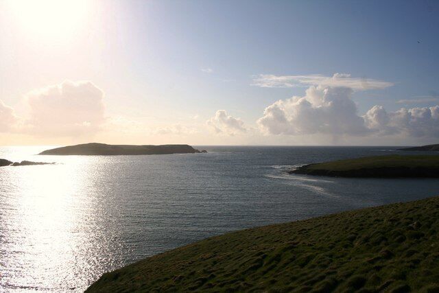 A coastal landscape with a grassy hill, calm sea, and a distant island under a clear sky.