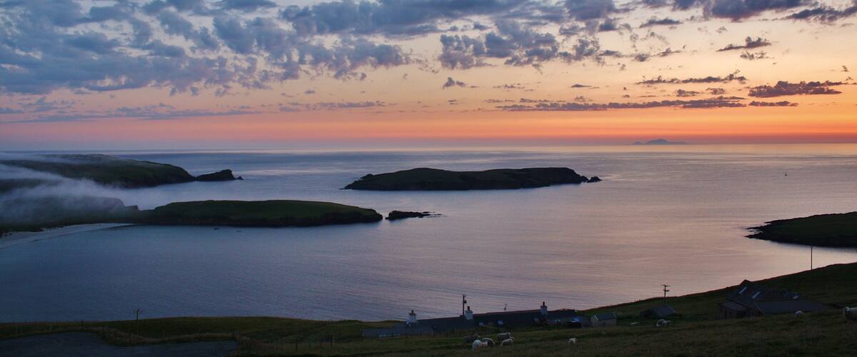 A coastal landscape with grassy hills, a calm sea, and a sunset sky with scattered clouds.