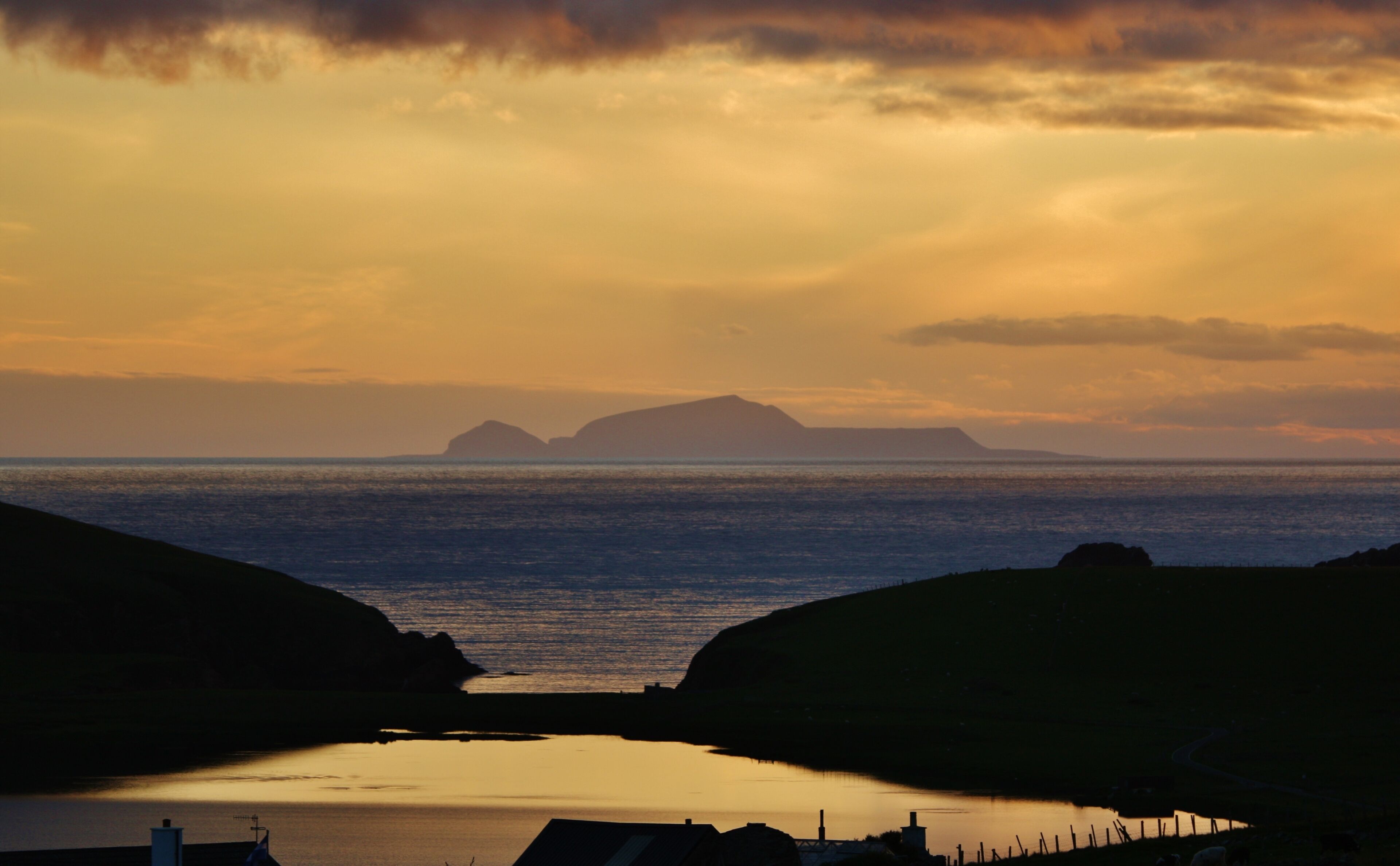 A coastal sunset with a calm sea, hills, and distant islands.