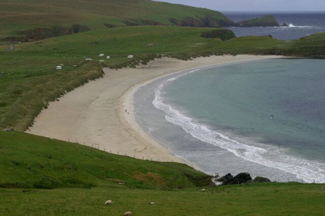 A coastal landscape with a sandy beach, grassy hills, and a rocky outcrop.