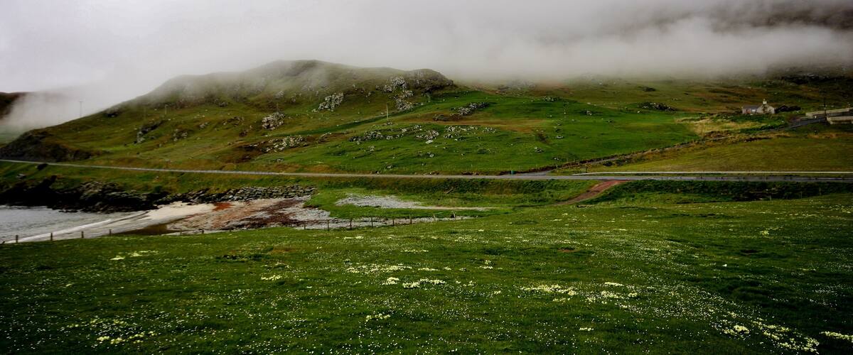 A coastal landscape with green fields, a rocky hill, and a bridge.