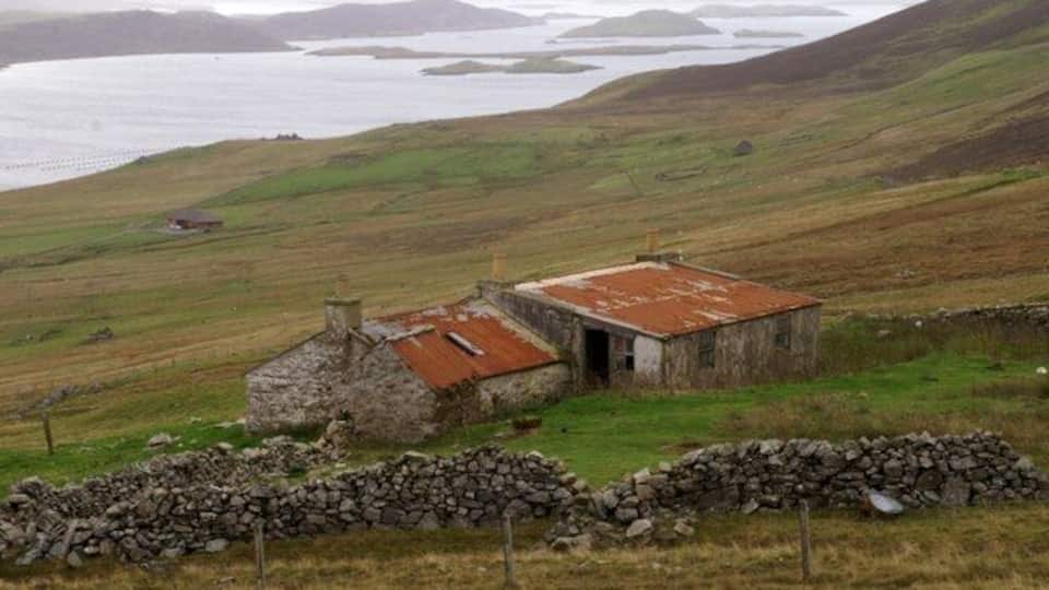 A stone house with a rusted roof in a grassy field with a stone wall in the foreground and a body of water in the background.