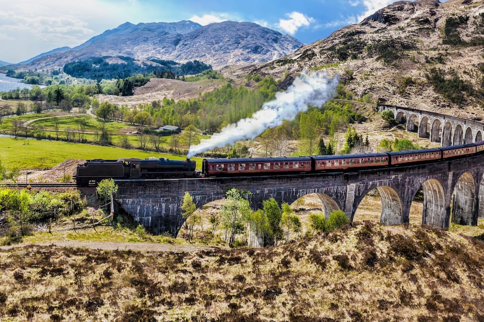 Um trem a vapor sobre um viaduto de pedra, cortando uma paisagem montanhosa com um lago ao fundo.