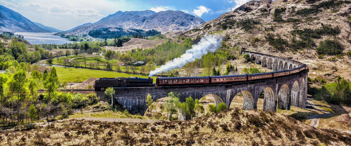 A steam train on a stone viaduct through a mountainous landscape with a lake in the background.