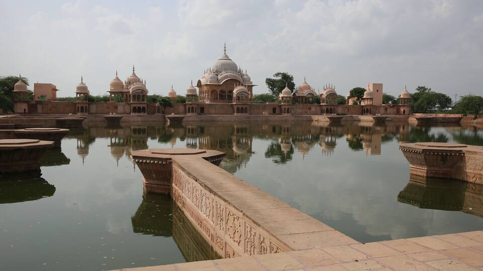 A historic building complex with domed structures and a calm reflective water body in the foreground.