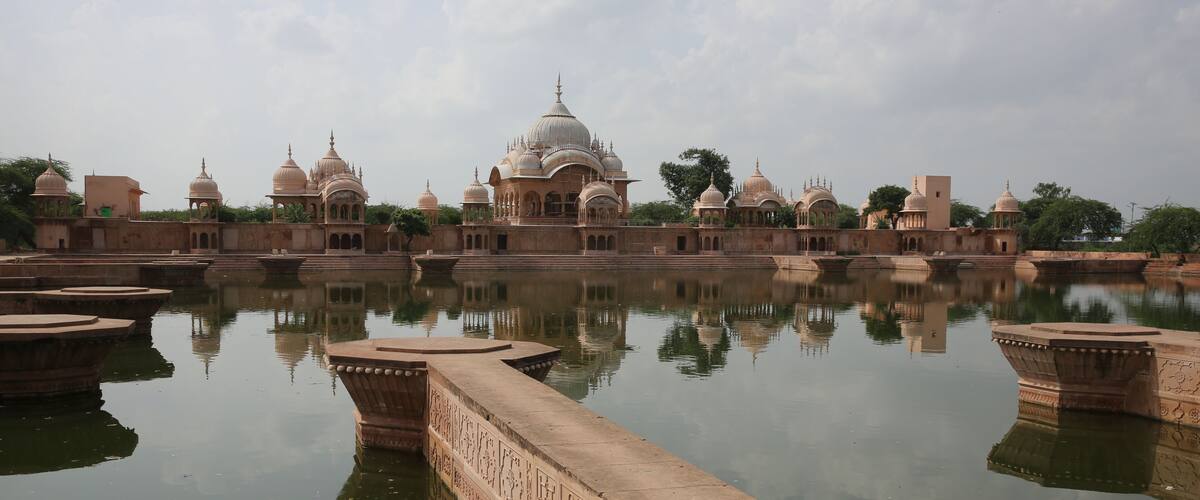A historic building complex with domed structures and a calm reflective water body in the foreground.