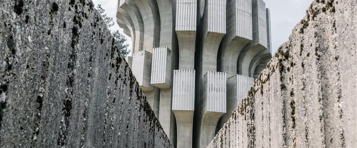 A modern architectural structure with a unique, ribbed design, flanked by concrete walls with holes, leading towards a clear sky.