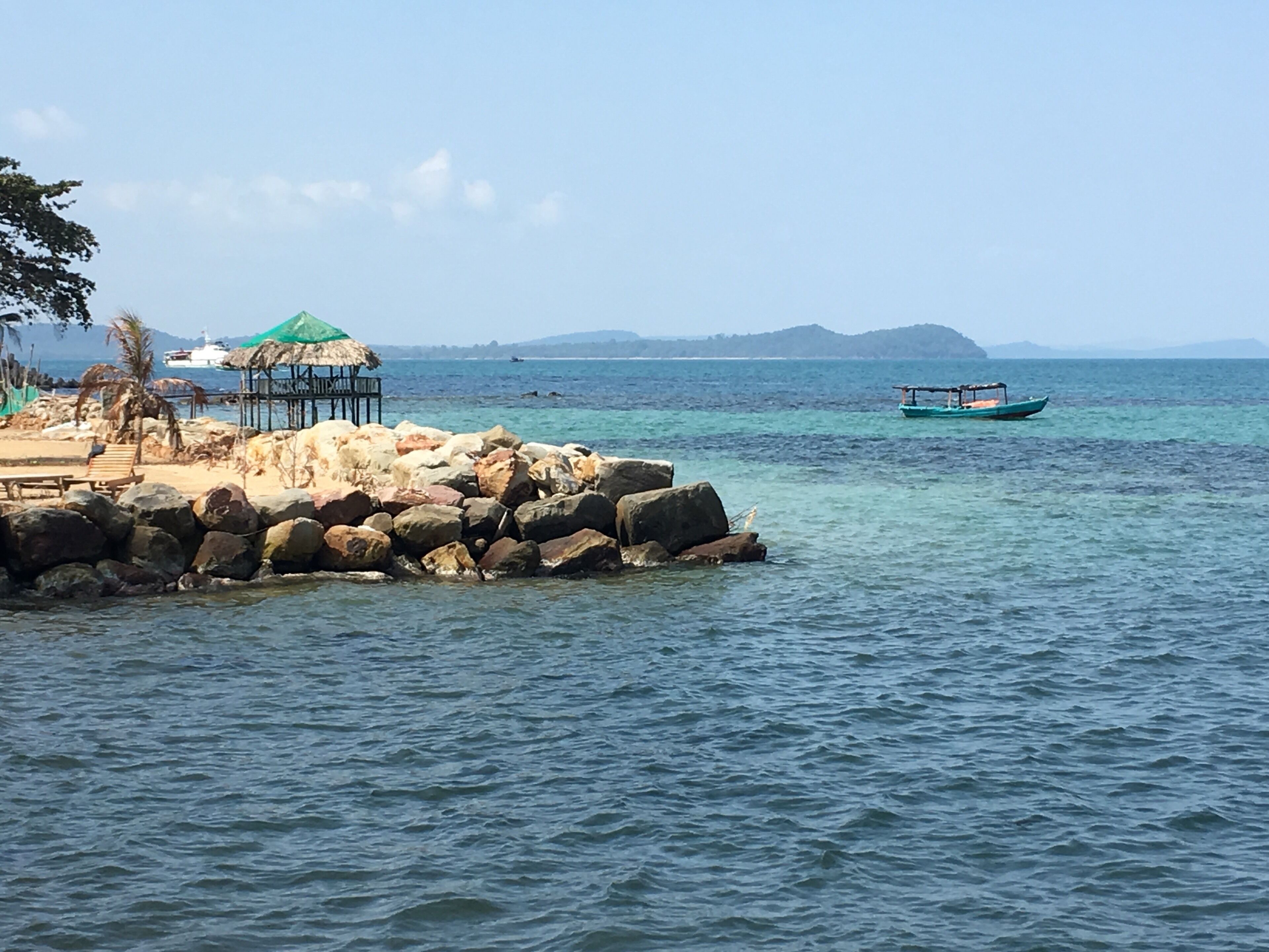 A rocky shoreline with a thatched-roof hut, a wooden bench, and a small boat in the water.