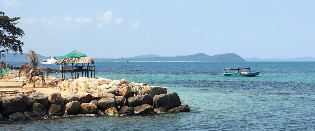 A rocky shoreline with a thatched-roof hut, a wooden bench, and a small boat in the water.