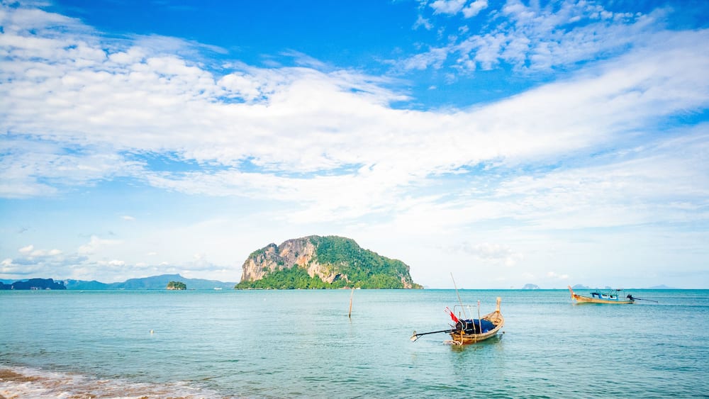 A calm sea with a small boat anchored near the shore, a large green island in the distance, and a clear blue sky with scattered clouds.