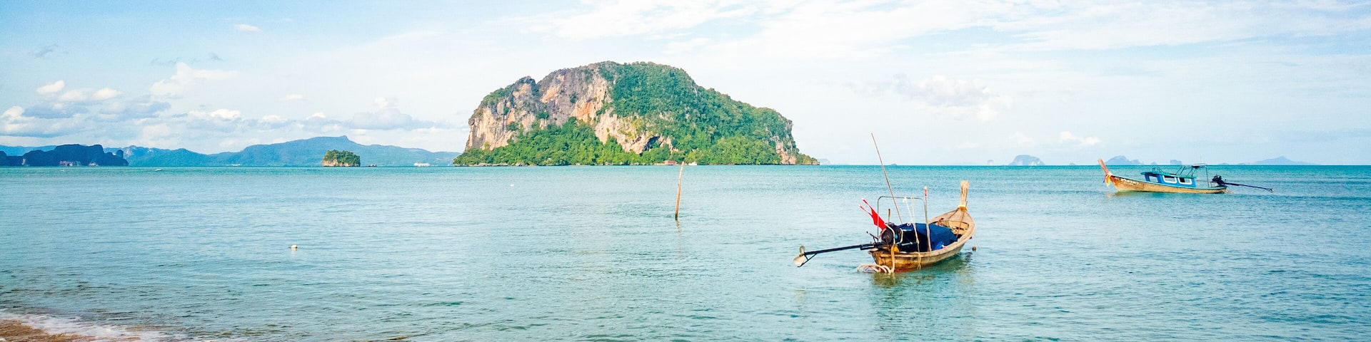 A calm sea with a small boat anchored near the shore, a large green island in the distance, and a clear blue sky with scattered clouds.