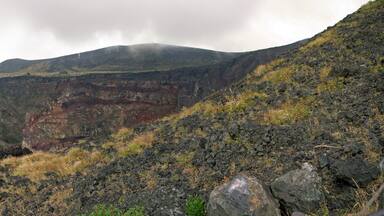 A volcanic landscape with a prominent crater, grassy slopes, and a cloudy sky.