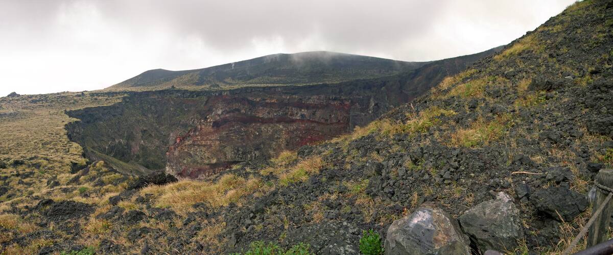 A volcanic landscape with a prominent crater, grassy slopes, and a cloudy sky.