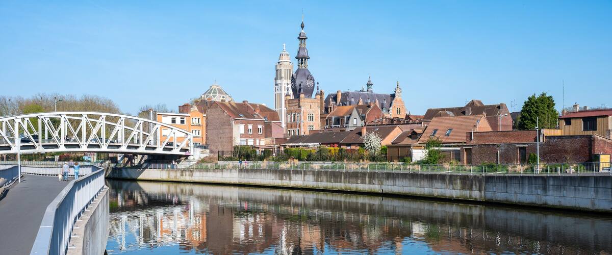 A bridge over a calm river reflecting buildings and a tower.