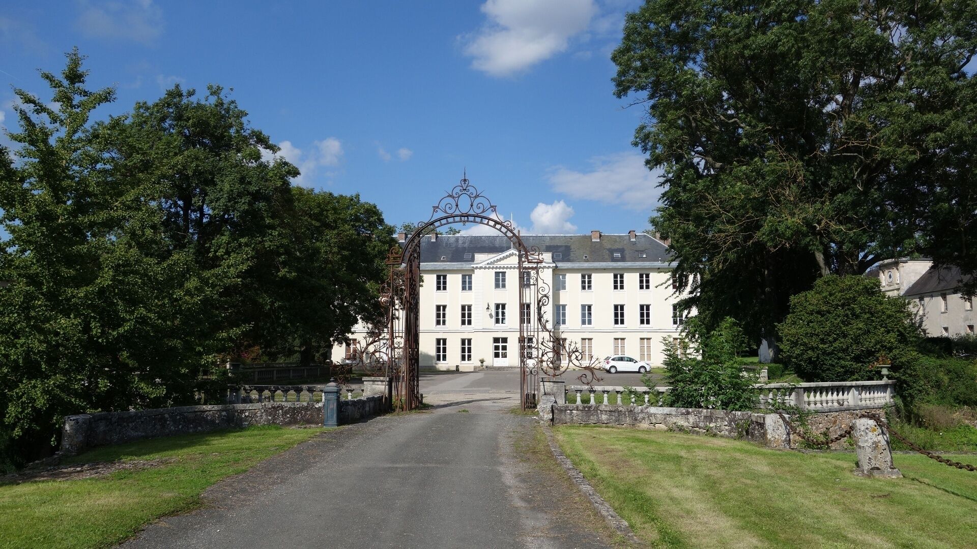 A gated entrance to a large, white building with a symmetrical facade, surrounded by lush greenery and a clear blue sky.