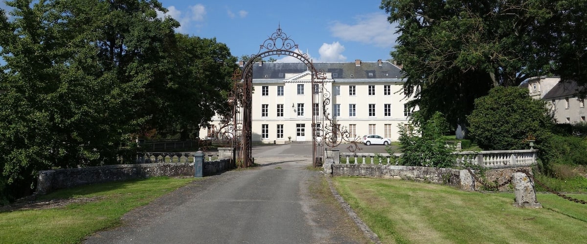 A gated entrance to a large, white building with a symmetrical facade, surrounded by lush greenery and a clear blue sky.