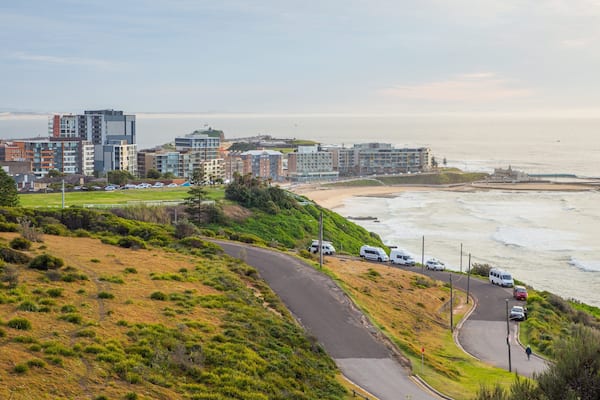 A coastal road with vehicles, a beach, and residential buildings.