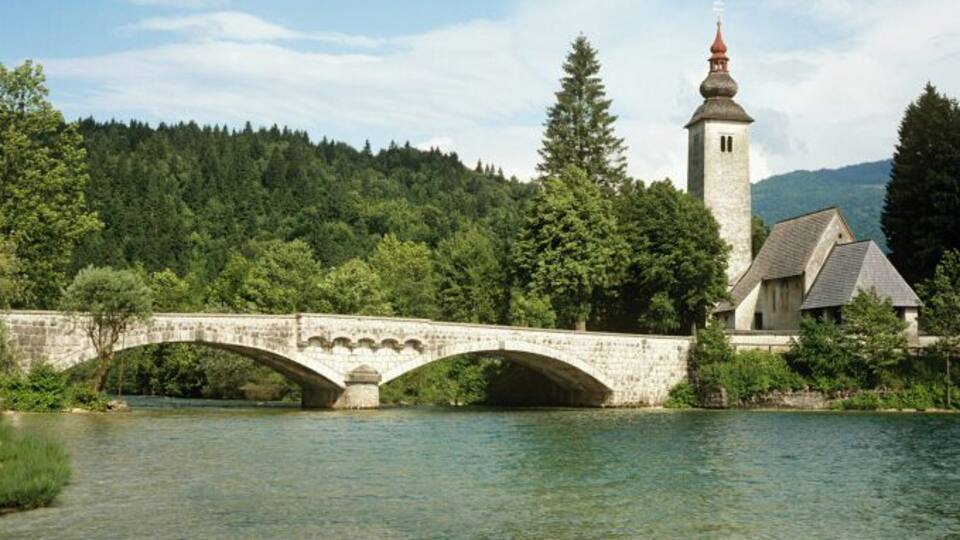 A stone bridge over a river, a church with a tower, and a forested hillside.
