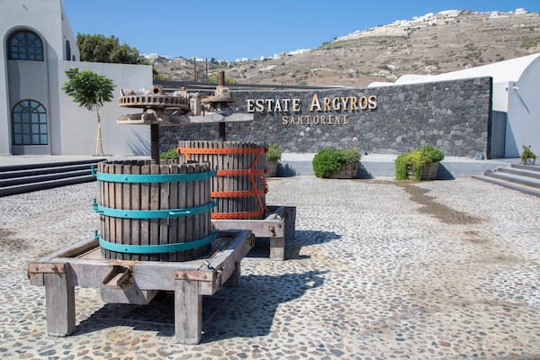 Un mur de pierre sur lequel on peut lire l’inscription « ESTATE ARGYROS SANTORINI », et un chariot en bois en forme de baril, posé sur un sol pavé.