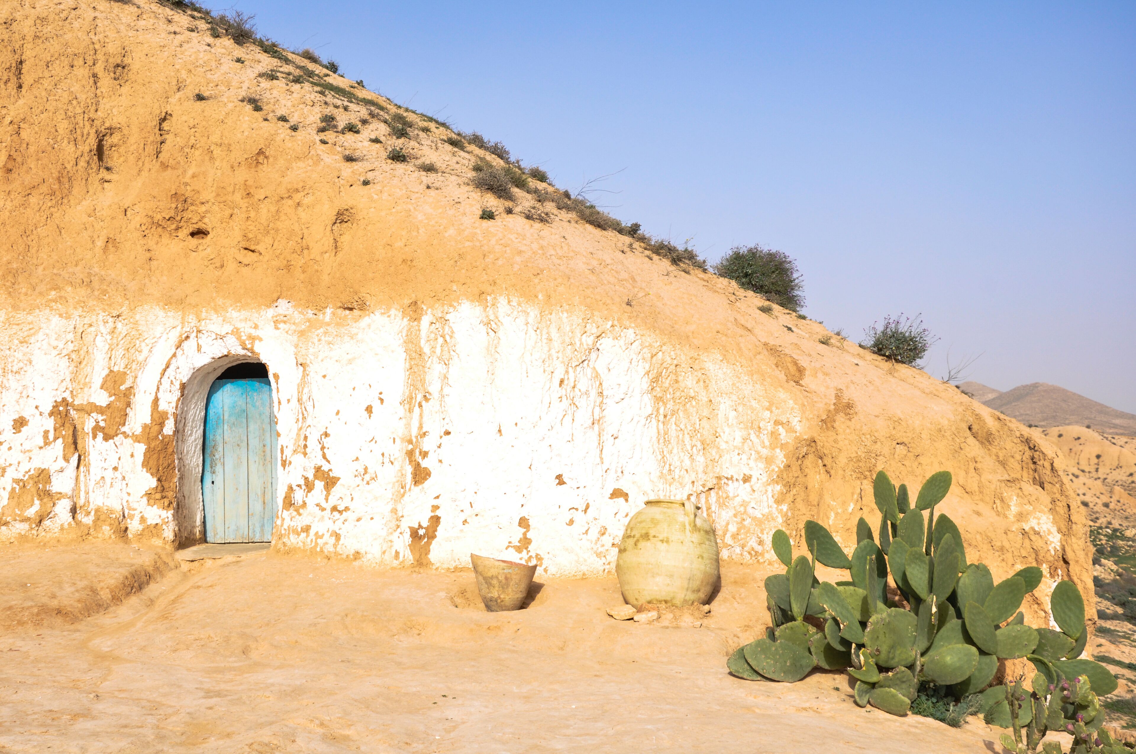 A desert landscape with a traditional mud-brick house, a blue door, a large pot, and a cactus.