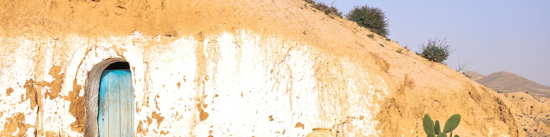 A desert landscape with a traditional mud-brick house, a blue door, a large pot, and a cactus.