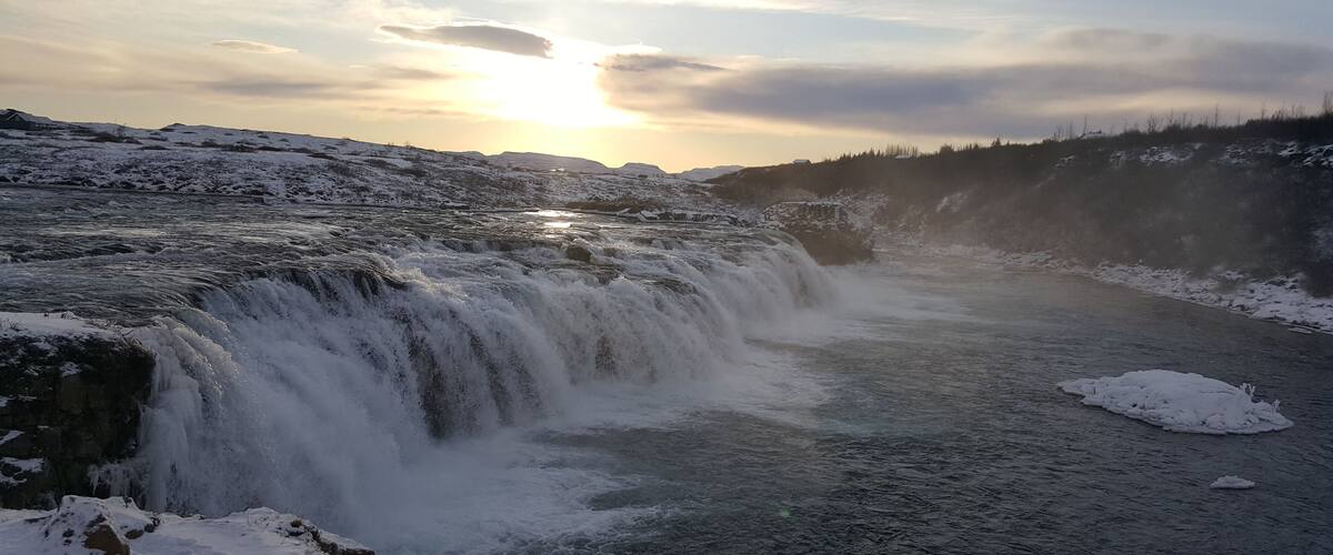 A waterfall cascading over snow-covered rocks with a sunset in the background.