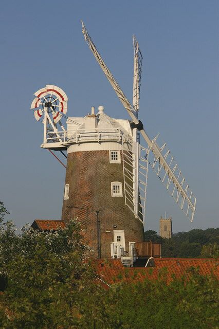 A windmill with a white windlass and a brick base, surrounded by greenery and a red-roofed building.