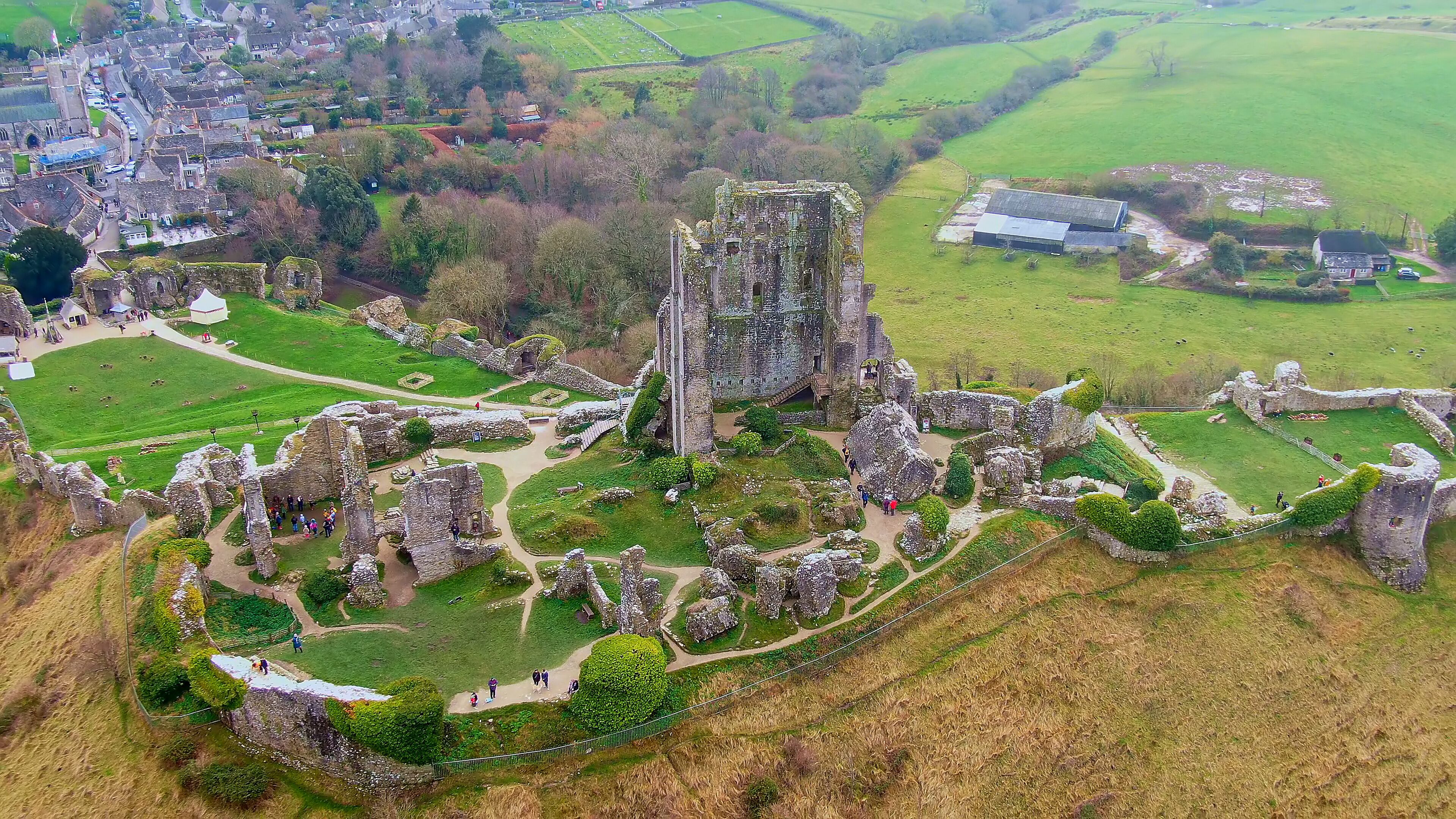 An aerial view of a historic castle ruins with surrounding green fields and a small village in the background.
