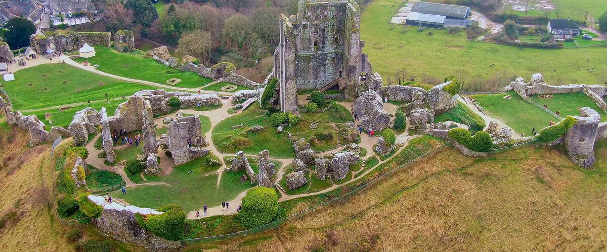 An aerial view of a historic castle ruins with surrounding green fields and a small village in the background.