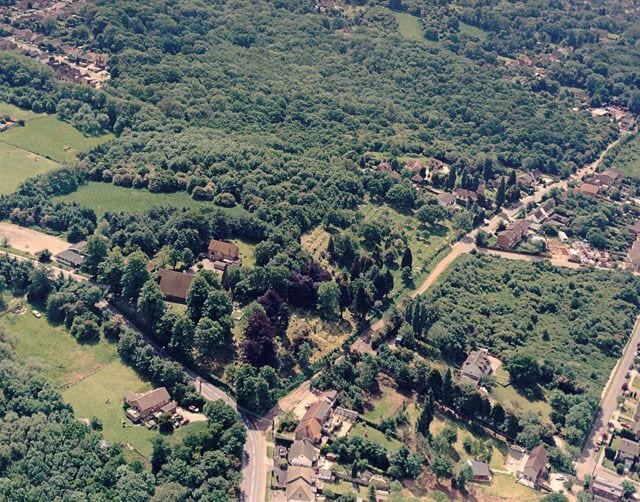 Aerial view of St Peters Church and Coombe Wood, Thundersley. St Peter's Church lies below-left of centre. The church spire is obscured by the trees. The road skirting the lower left is the Church Road. The road extending off this towards the upper right id Rhoda Road North. Much of this is unmade. The wood occupying the upper part is Coombe Wood. The lower right of the image shows the escarpment, seen from the other direction in 1635727.