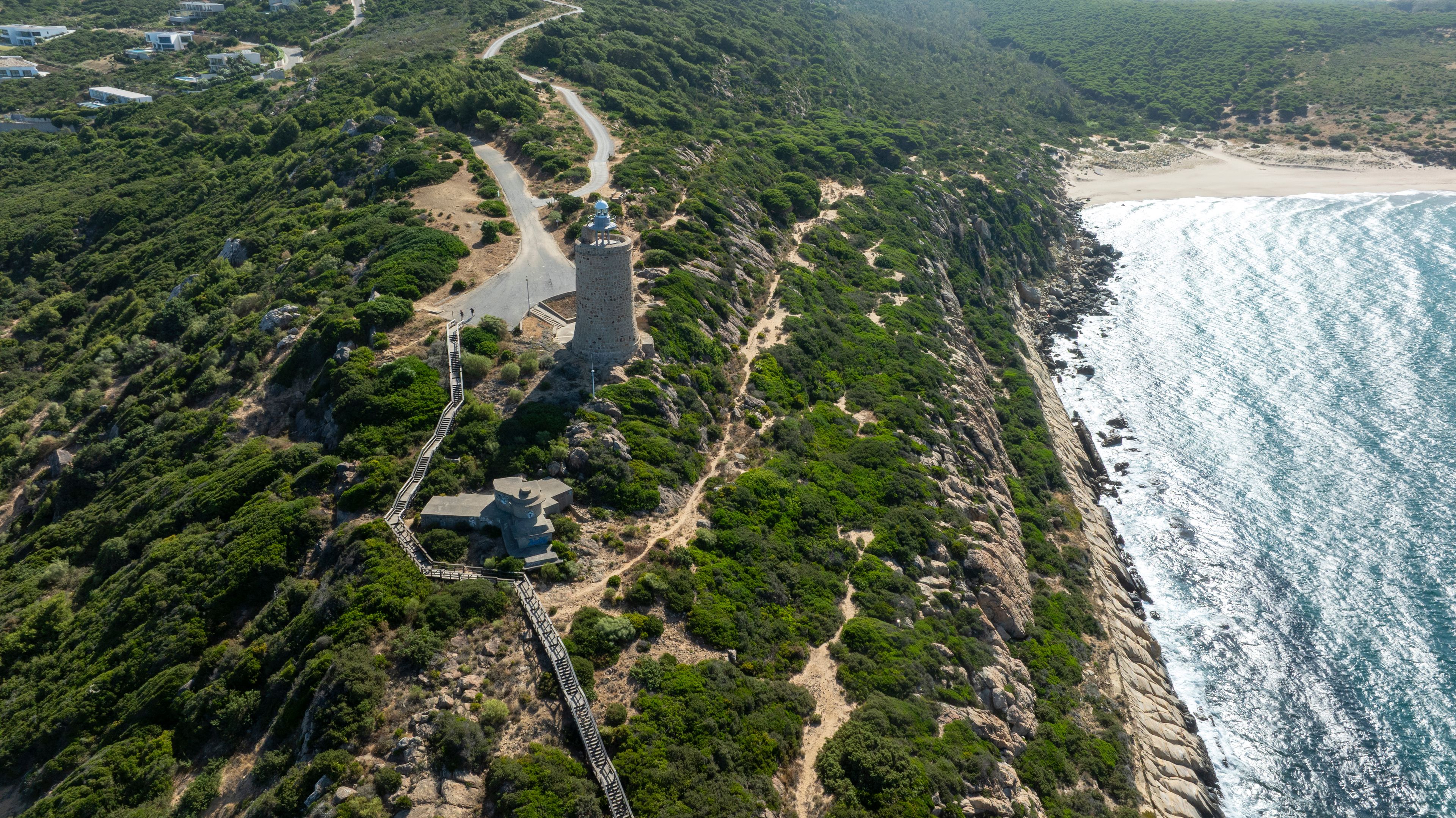 An aerial view of a coastal area with a winding road, a tower, and a beach.